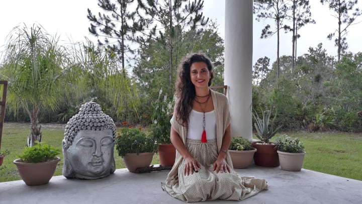 An image of Dass Shanti kneeling on a porch next to a Buddha with trees in the background.